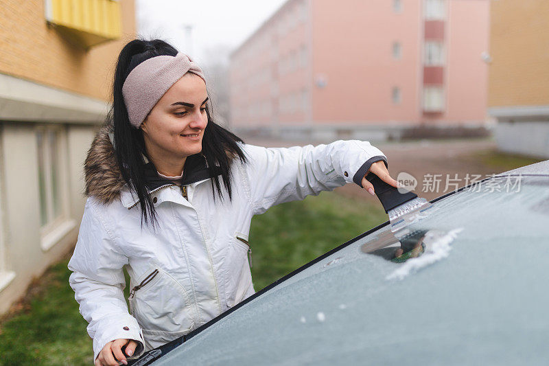 美丽的年轻女子在早上刮掉她汽车挡风玻璃上的冰/霜